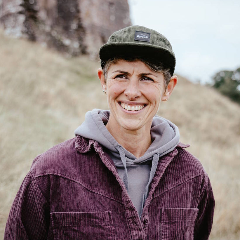 A smiling woman outdoors wearing an olive green Bunnet cap, a gray hoodie, and a purple corduroy overshirt, with a rocky hillside in the background.