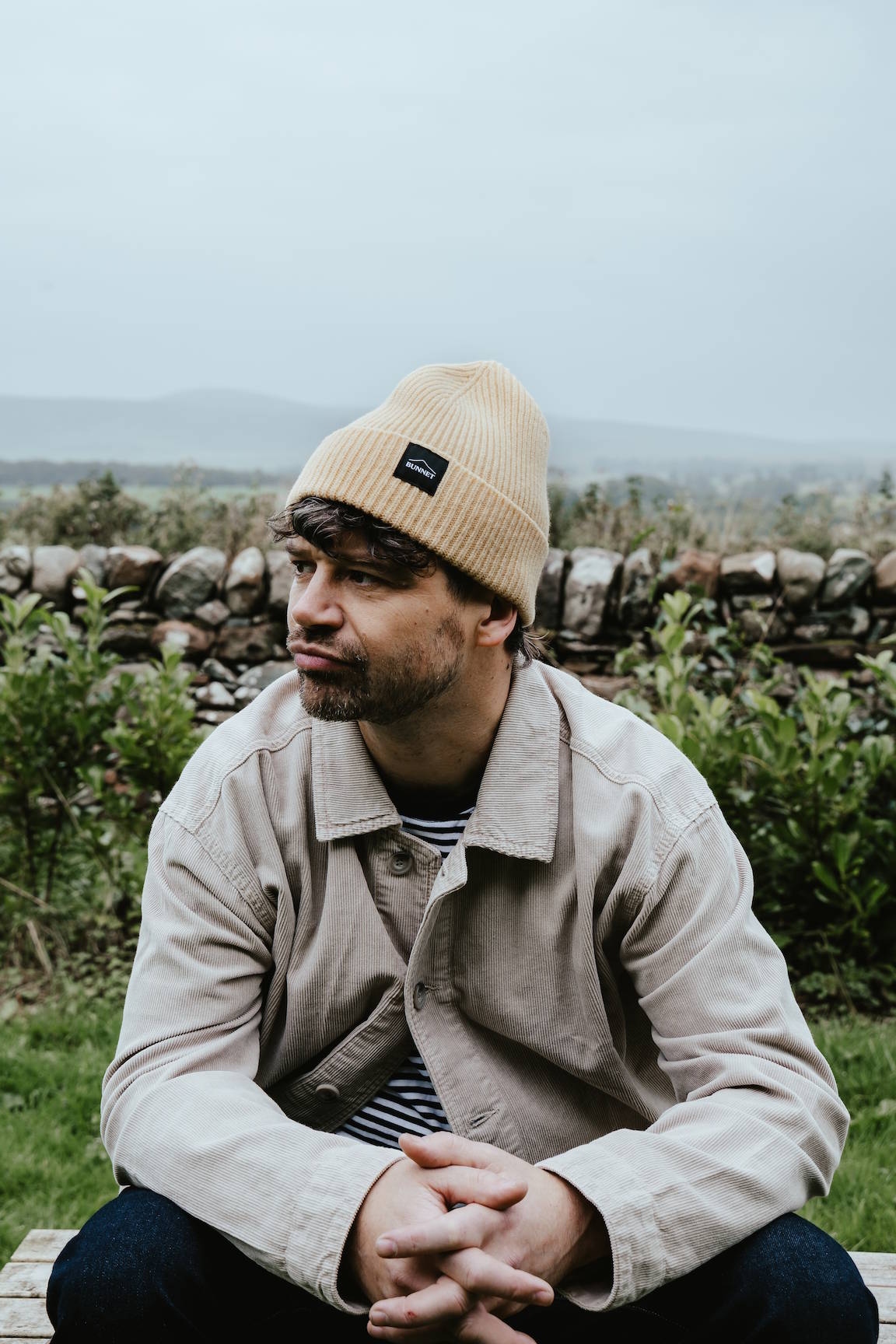 A man sitting outdoors on a bench wearing a beige knit beanie with a small black Bunnet brand patch, a light beige overshirt, and a striped shirt, with a stone wall and greenery in the background.