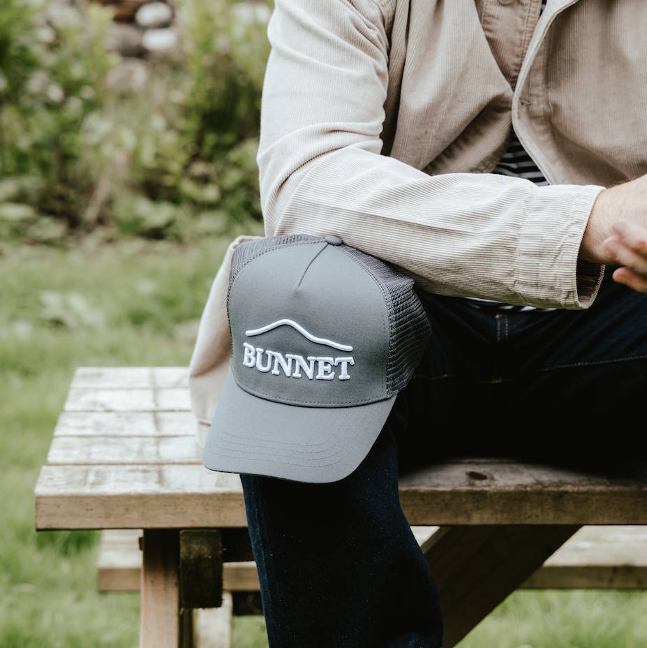 A person sitting outdoors on a wooden bench wearing a light beige shirt and dark pants, with a gray BUNNET trucker cap resting on their lap. The cap has a white embroidered mountain graphic and the word 'BUNNET' on the front.