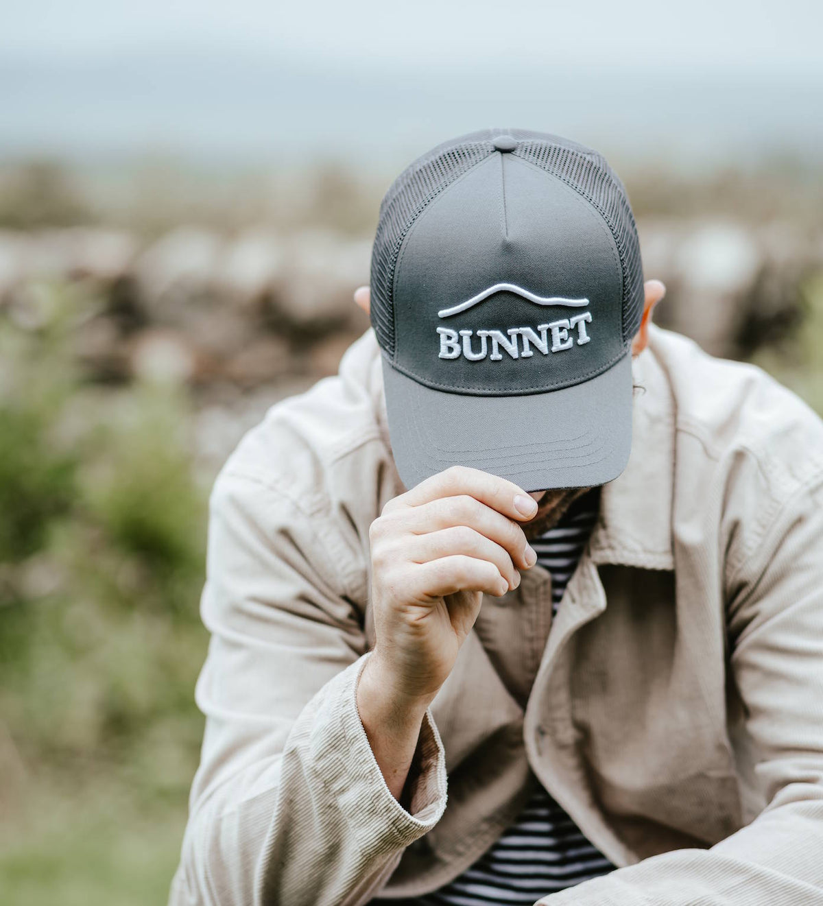 A man wearing a dark gray trucker hat with 'BUNNET' embroidered in white on the front, sitting outdoors in a beige jacket over a striped shirt.