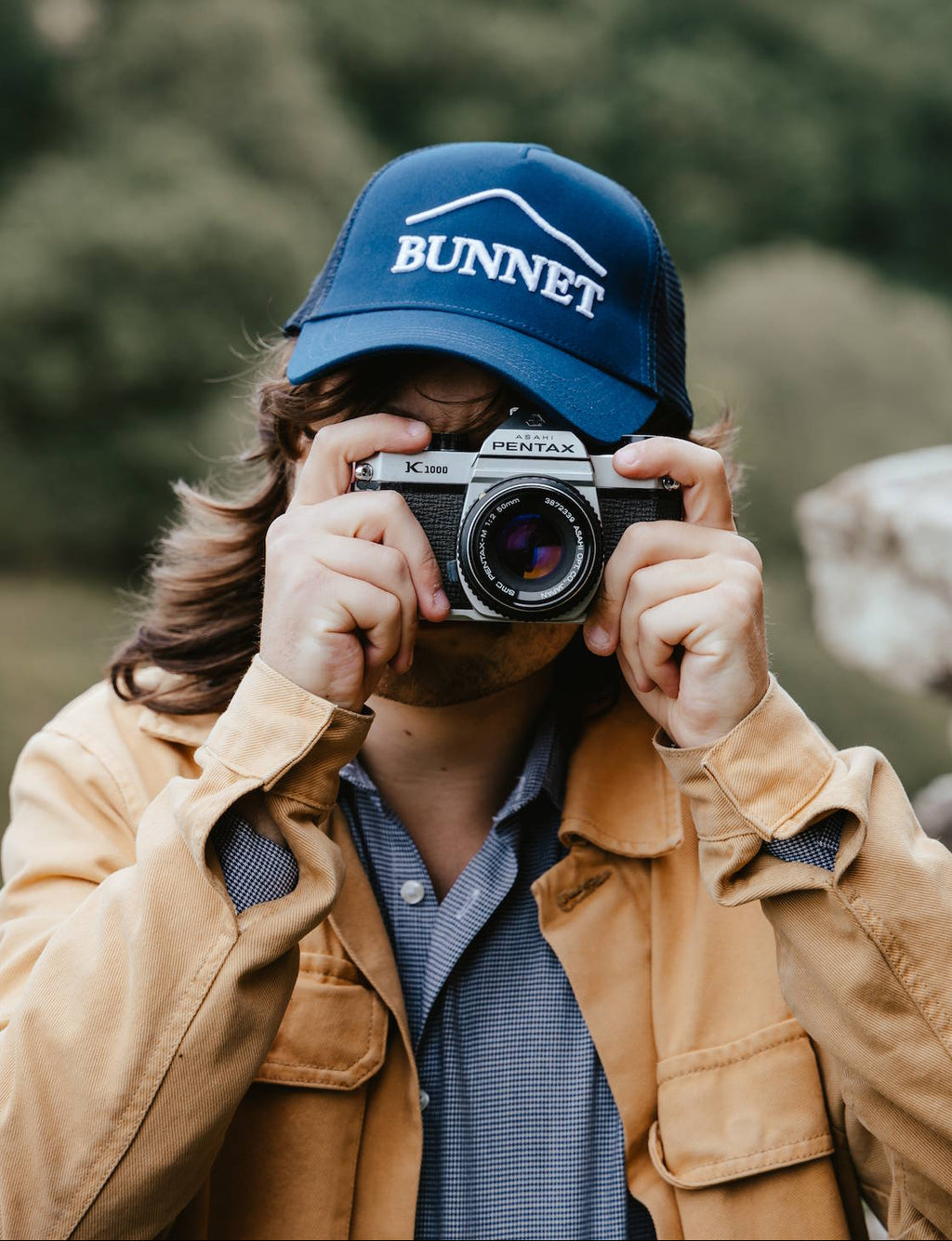 A person wearing a blue 'BUNNET' trucker hat and a tan jacket holds a vintage camera to their face, with a blurred outdoor background of trees and rocks.