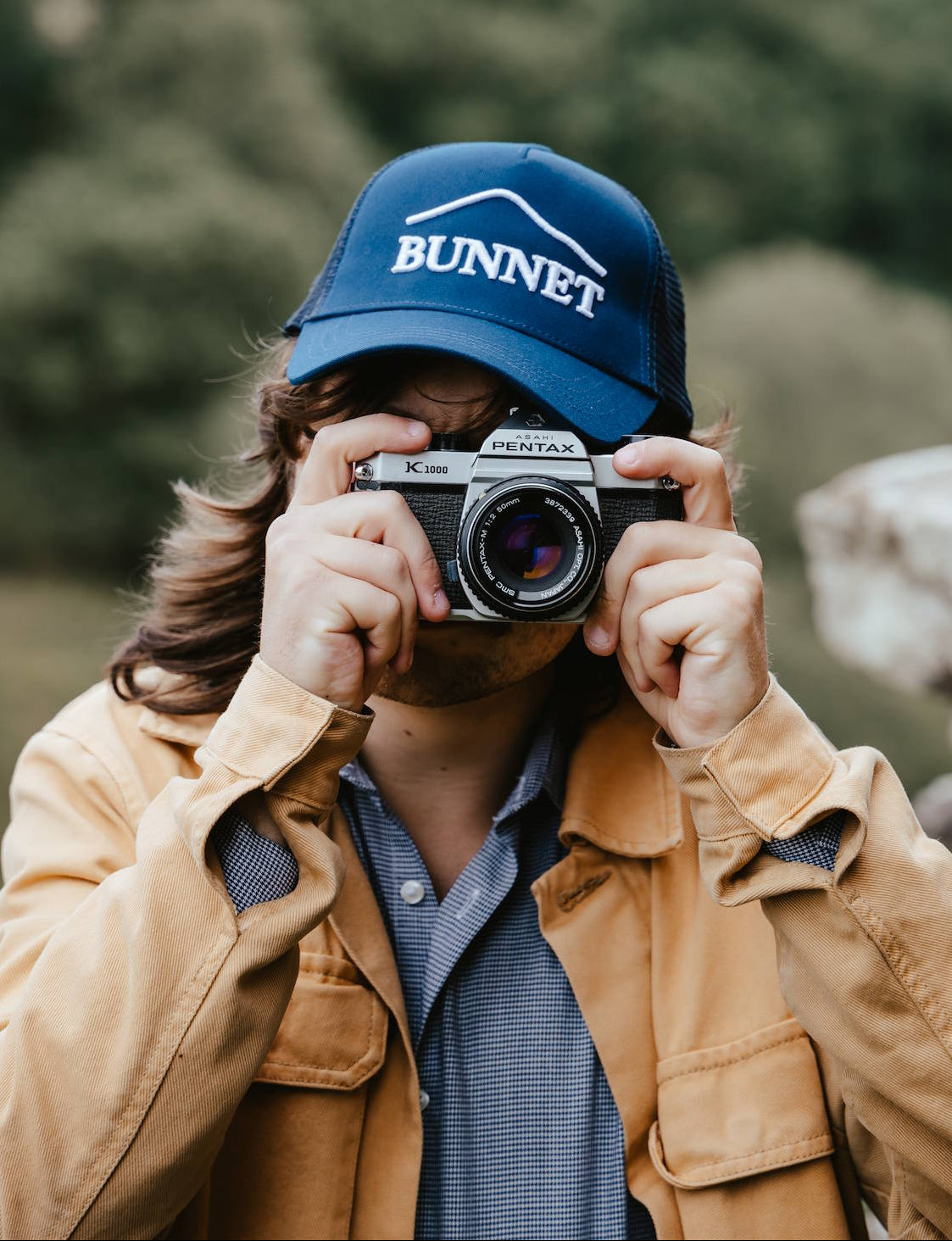 A person wearing a blue 'BUNNET' trucker hat and a tan jacket holds a vintage camera to their face, with a blurred outdoor background of trees and rocks.