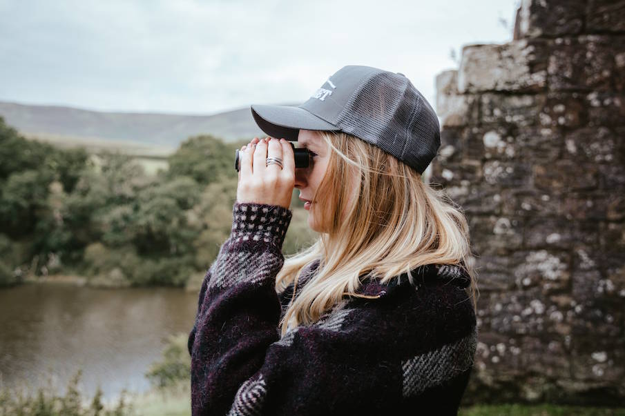 A woman in a dark gray Bunnet trucker hat and a dark patterned sweater looks through binoculars outdoors near a stone wall and water.