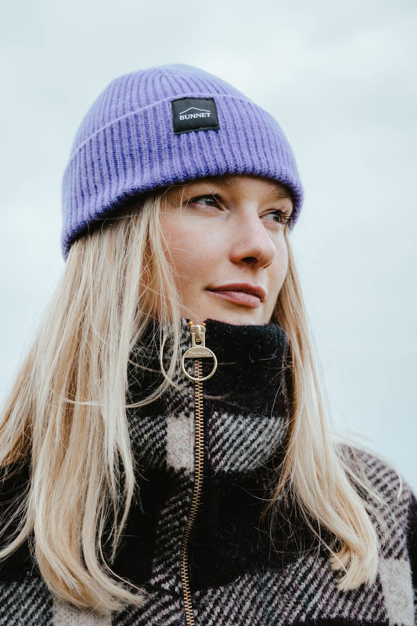 A woman wearing a lavender-purple ribbed knit beanie with a black rectangular BUNNET brand patch on the front, paired with a black and gray plaid zip-up coat against an overcast sky.