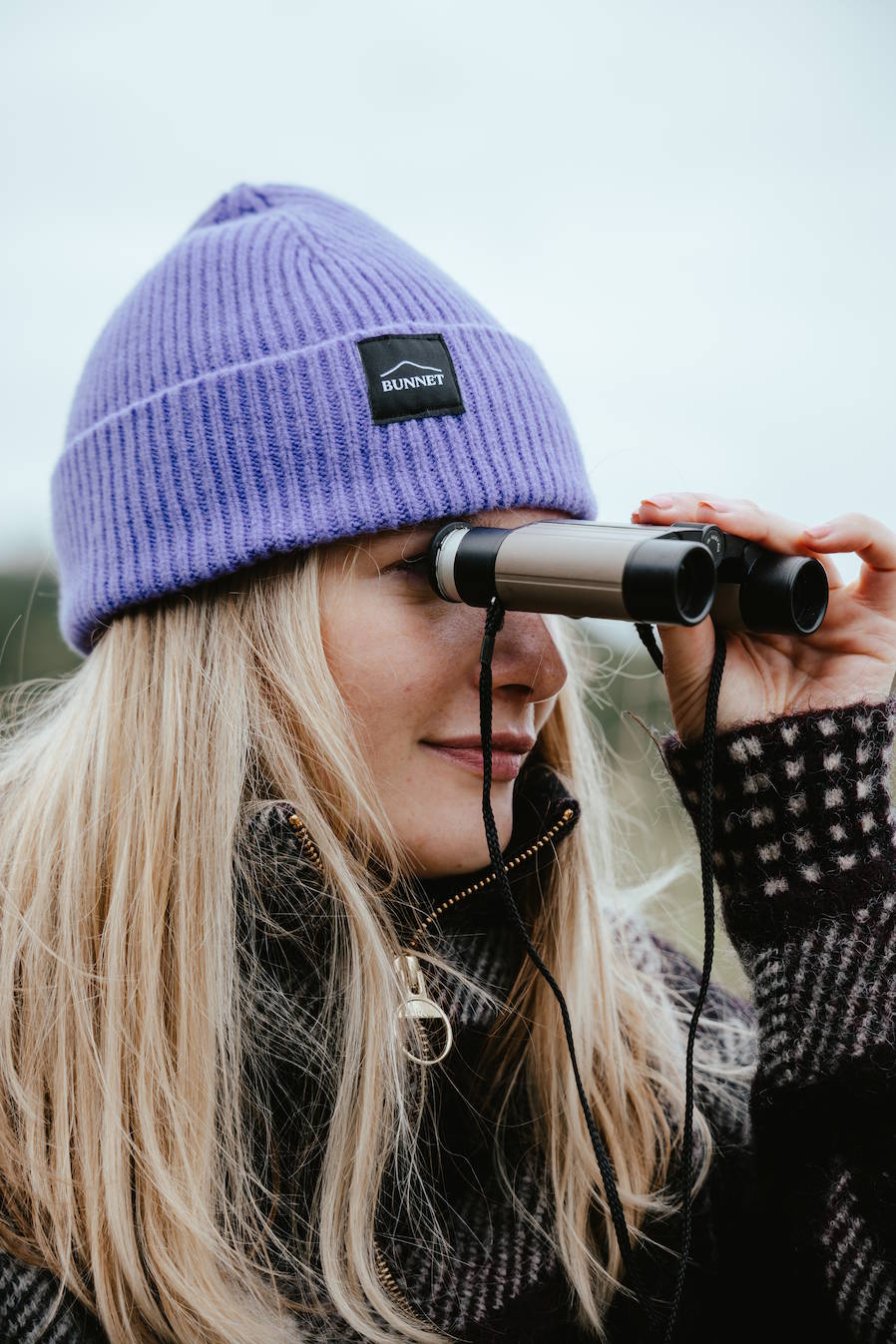 A woman wearing a lavender-purple ribbed knit BUNNET beanie with a black brand patch looks through binoculars outdoors.