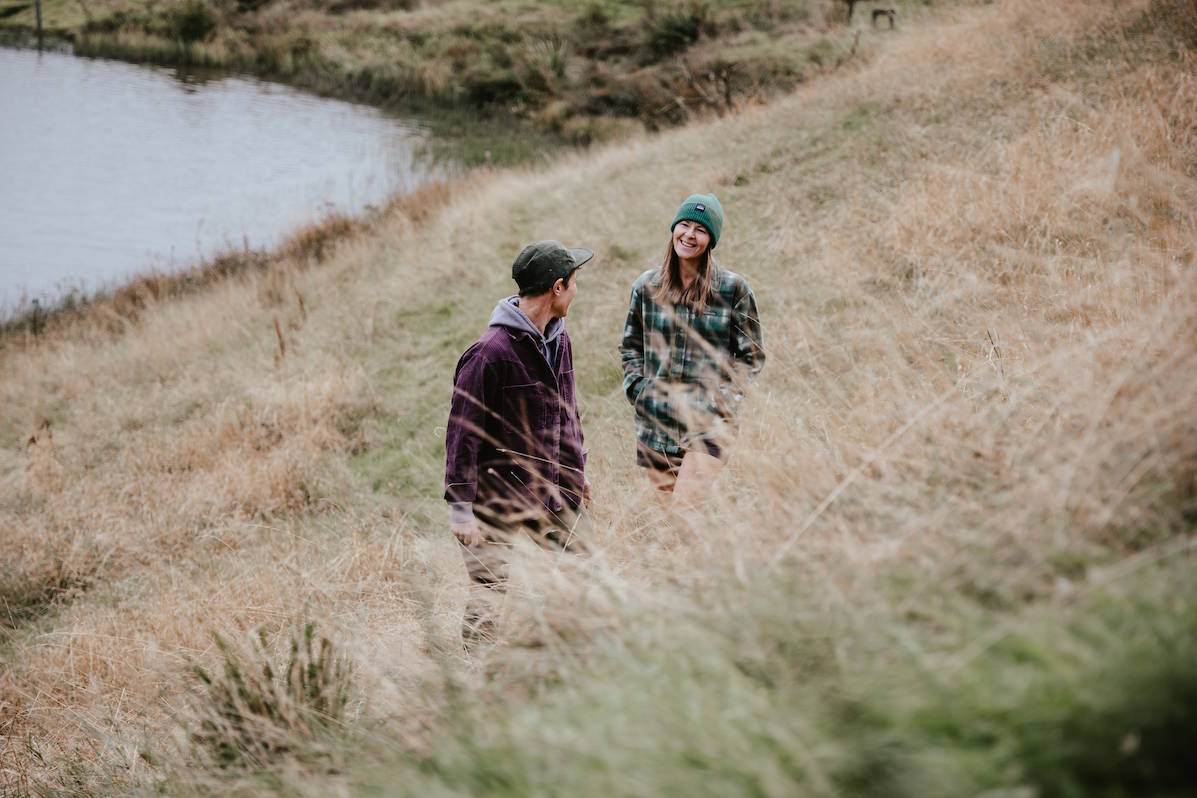 Two people walking through tall dry grass beside a body of water in an outdoor setting wearing bunnets