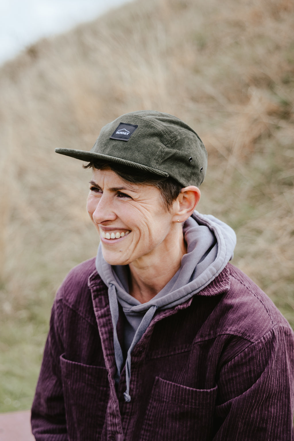 A smiling woman outdoors wearing an olive green corduroy cap with a small black rectangular BUNNET logo patch, a gray hoodie, and a dark purple corduroy jacket, with a blurred grassy hill in the background.