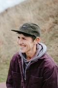 A smiling woman outdoors wearing an olive green corduroy cap with a small black rectangular BUNNET logo patch, a gray hoodie, and a dark purple corduroy jacket, with a blurred grassy hill in the background.