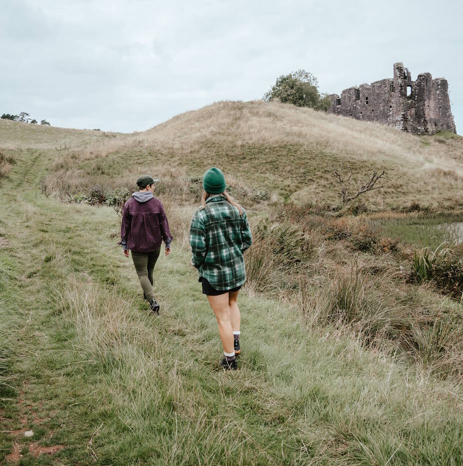 Two people walking away on a grassy hill toward a stone ruin under an overcast sky, with one wearing a green beanie and green plaid shirt and the other wearing a dark jacket and green pants.