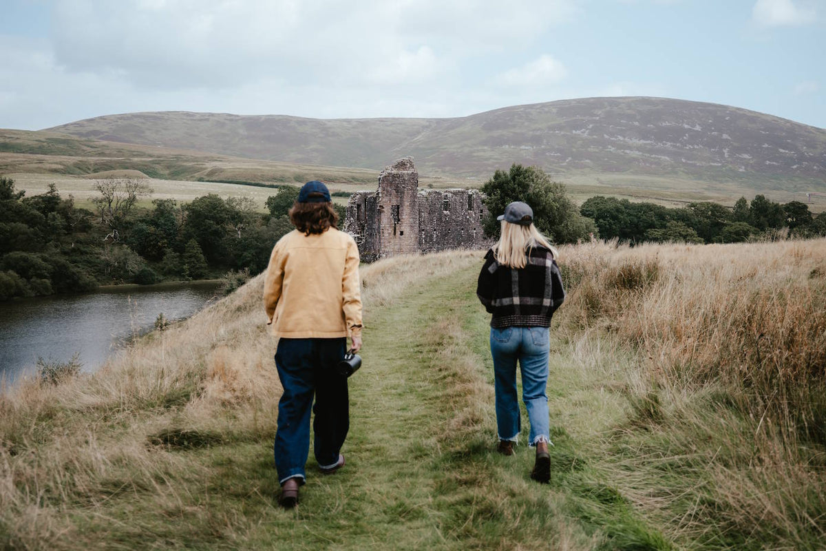 Two people walking away from the camera on a grassy path toward a stone ruin, with hills, trees, and a river in the background.