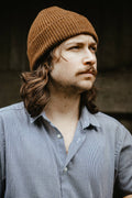 A man with long brown hair and a mustache wearing a brown ribbed knit BUNNET beanie and a blue-gray button-down shirt, shown in a close-up portrait.