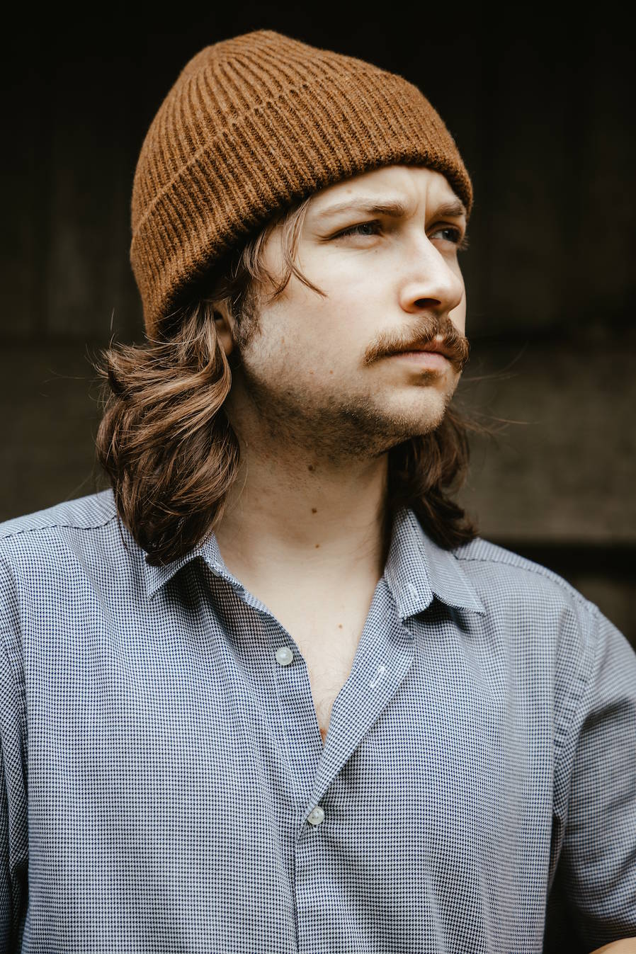A man with long brown hair and a mustache wearing a brown ribbed knit BUNNET beanie and a blue-gray button-down shirt, shown in a close-up portrait.