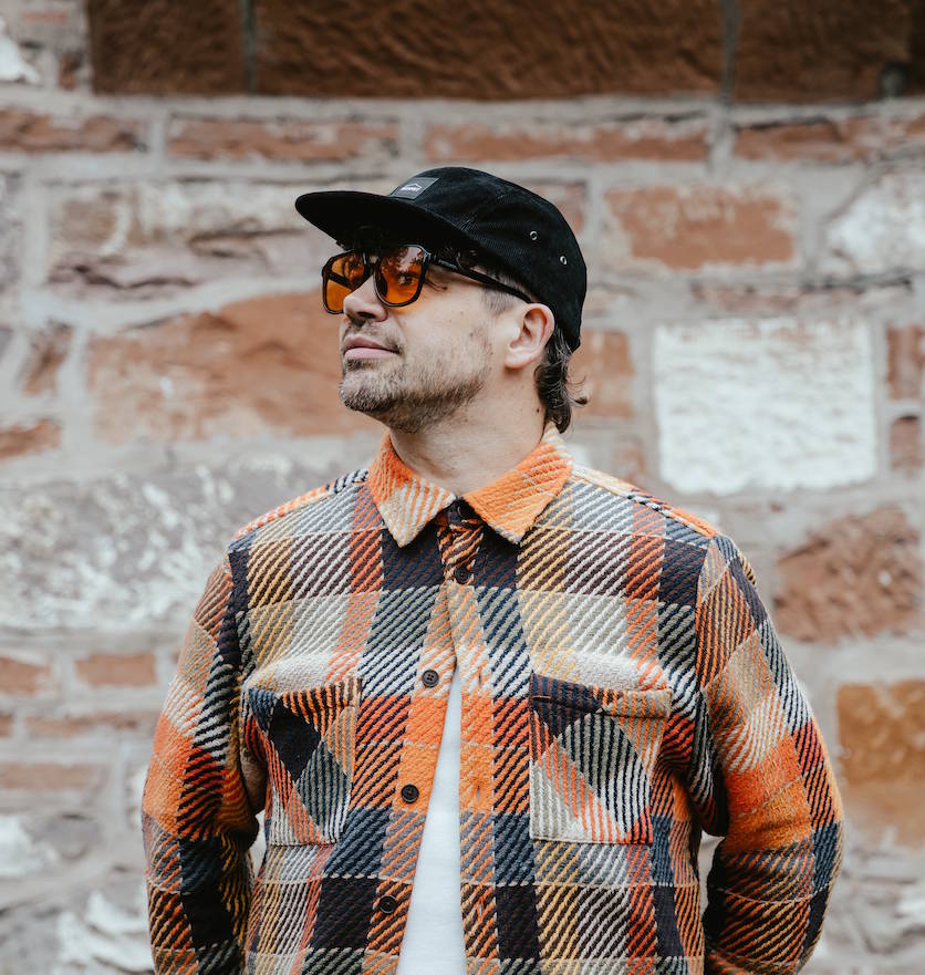 A man wearing a black Bunnet baseball cap, orange-tinted sunglasses, and a black, orange, and beige plaid button-up flannel shirt, standing in front of a rustic brick wall.
