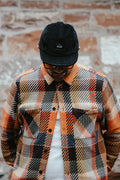 A man wearing a black corduroy Bunnet cap, orange-tinted sunglasses, and a black, orange, and beige plaid flannel shirt over a white undershirt, standing against a rustic brick wall.
