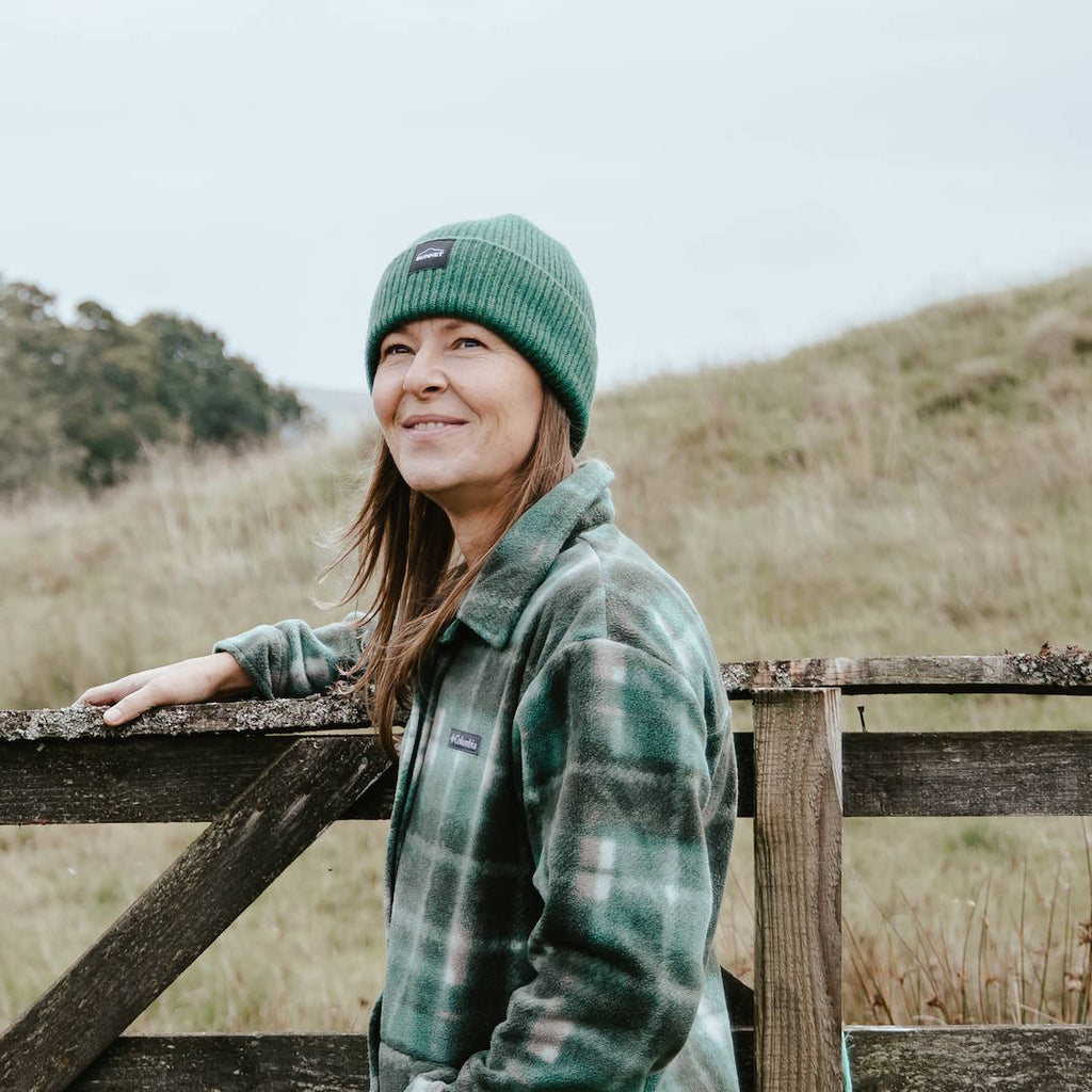 A woman wearing a green knit BUNNET beanie and a green plaid fleece jacket leans against a rustic wooden fence in an outdoor grassy field under an overcast sky.