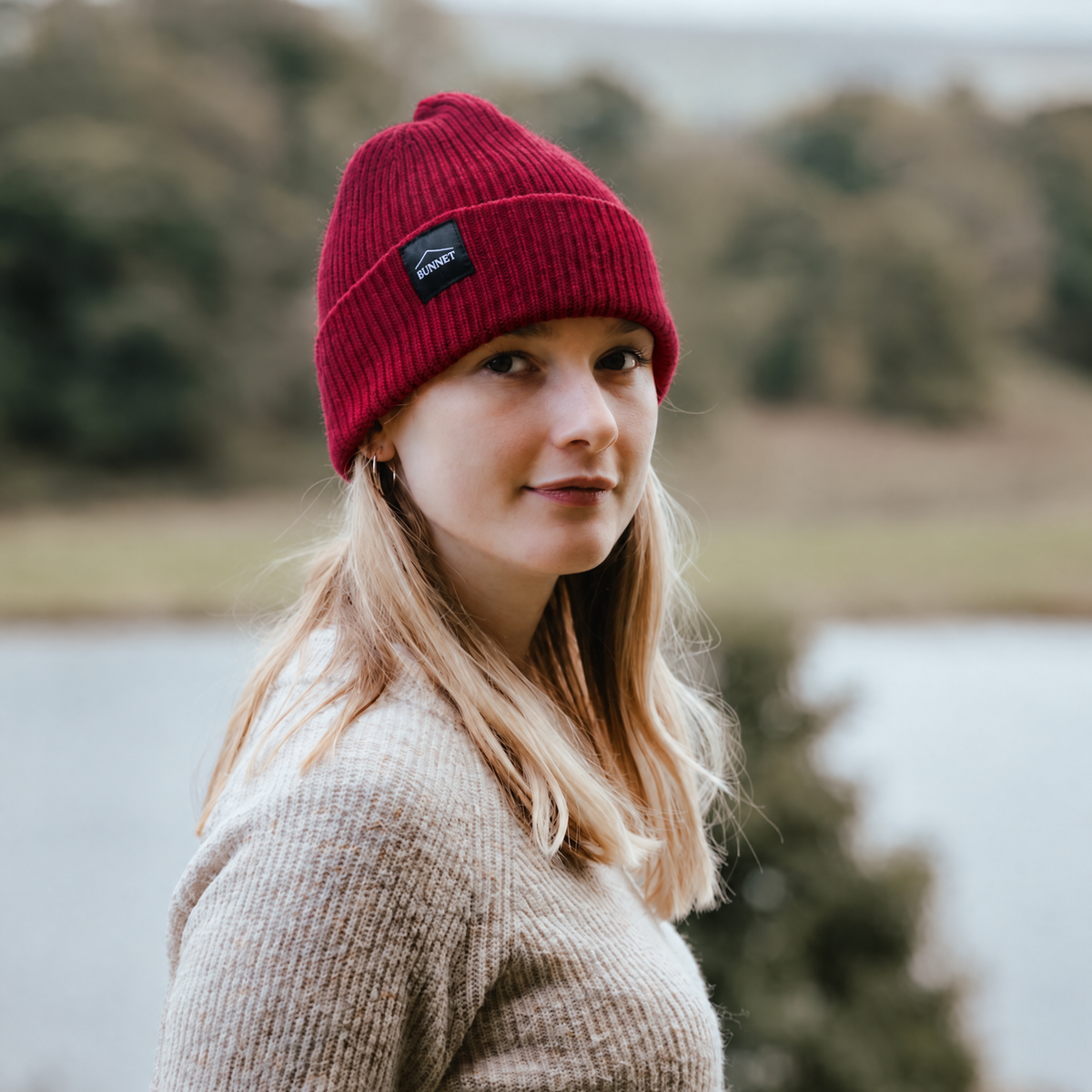 A woman wearing a red ribbed knit beanie with a small black brand Bunnet patch, standing outdoors in front of a blurred lake and trees.