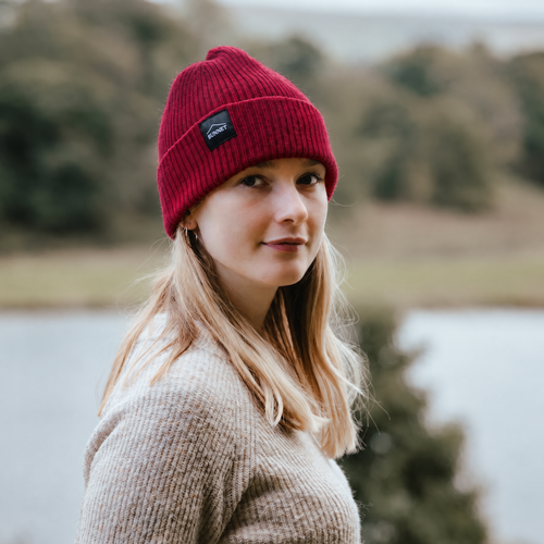 A woman wearing a red ribbed knit beanie with a small black brand Bunnet patch, standing outdoors in front of a blurred lake and trees.