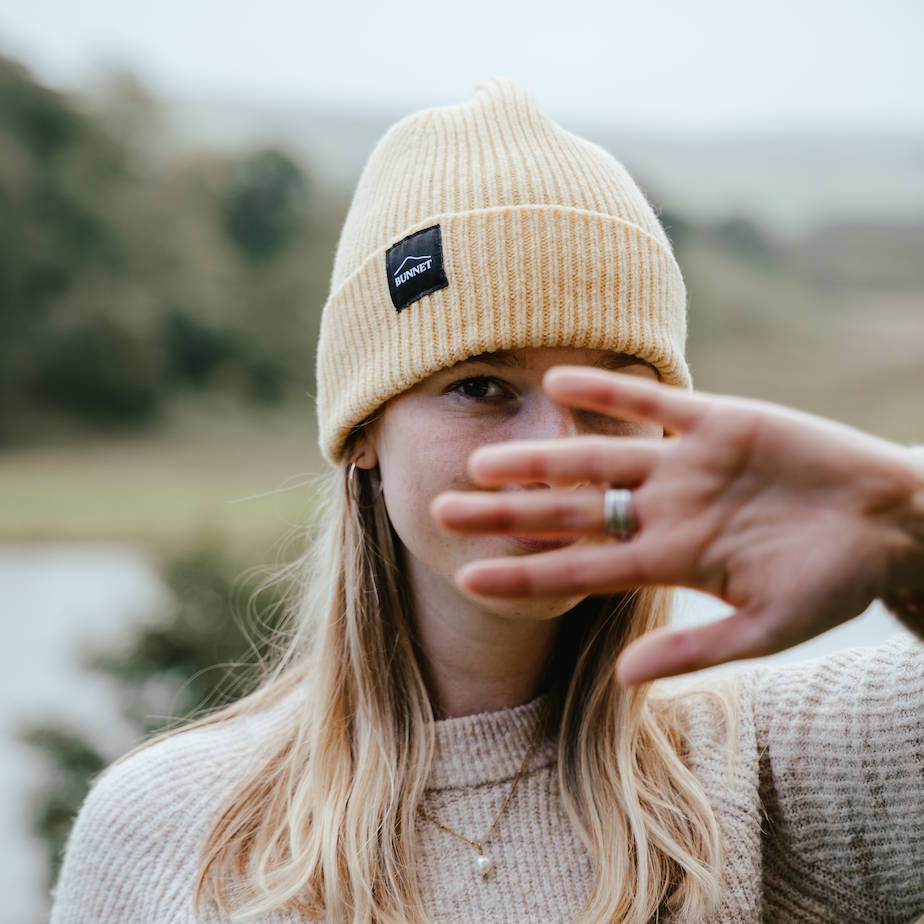 A woman wearing a Bunnet beige ribbed knit beanie with a black rectangular logo patch on the front, photographed outdoors with a blurred natural background. Her hand is raised in front of her face.