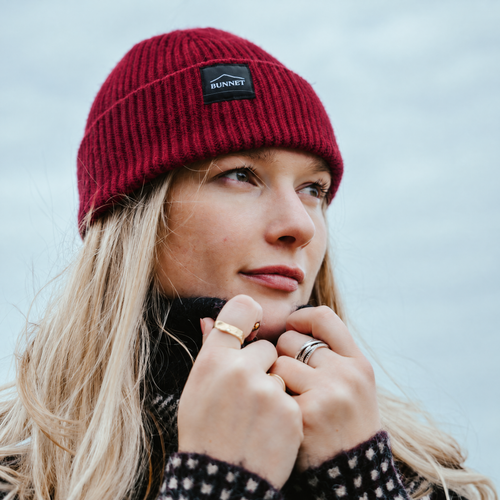 A woman wearing a burgundy ribbed knit beanie with a black rectangular patch that reads 'BUNNET' in white text, photographed outdoors against a soft gray sky.