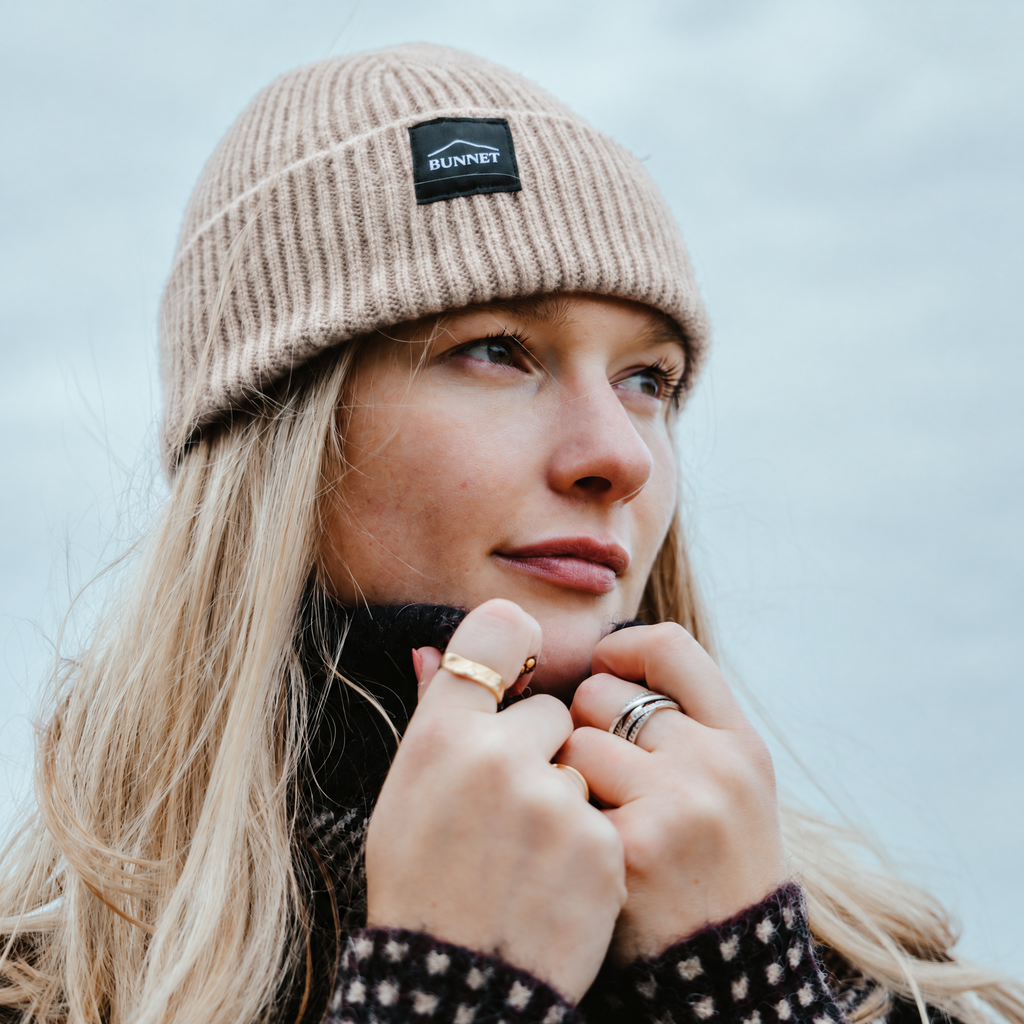 A close-up lifestyle shot of a woman wearing a beige ribbed knit beanie with a black 'BUNNET' logo patch on the front, set against a soft, overcast sky.