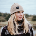 A woman outdoors wearing a beige ribbed knit beanie with a black rectangular logo patch that reads 'Bunnet'. She has long blonde hair and is wearing a dark patterned knit sweater.