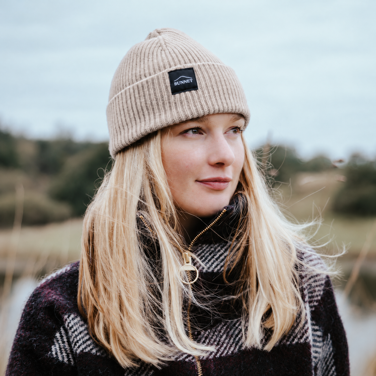 A woman outdoors wearing a beige ribbed knit beanie with a black rectangular logo patch that reads 'Bunnet'. She has long blonde hair and is wearing a dark patterned knit sweater.